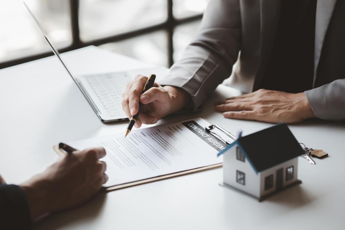 Closeup on the hands of two people holding pens, pointing at a contract on a desk with a laptop and a small model house.