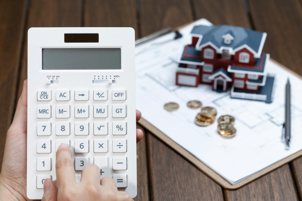 A hand operating a calculator in front of a house model on top of blueprints with scattered coins and a pen next to it.