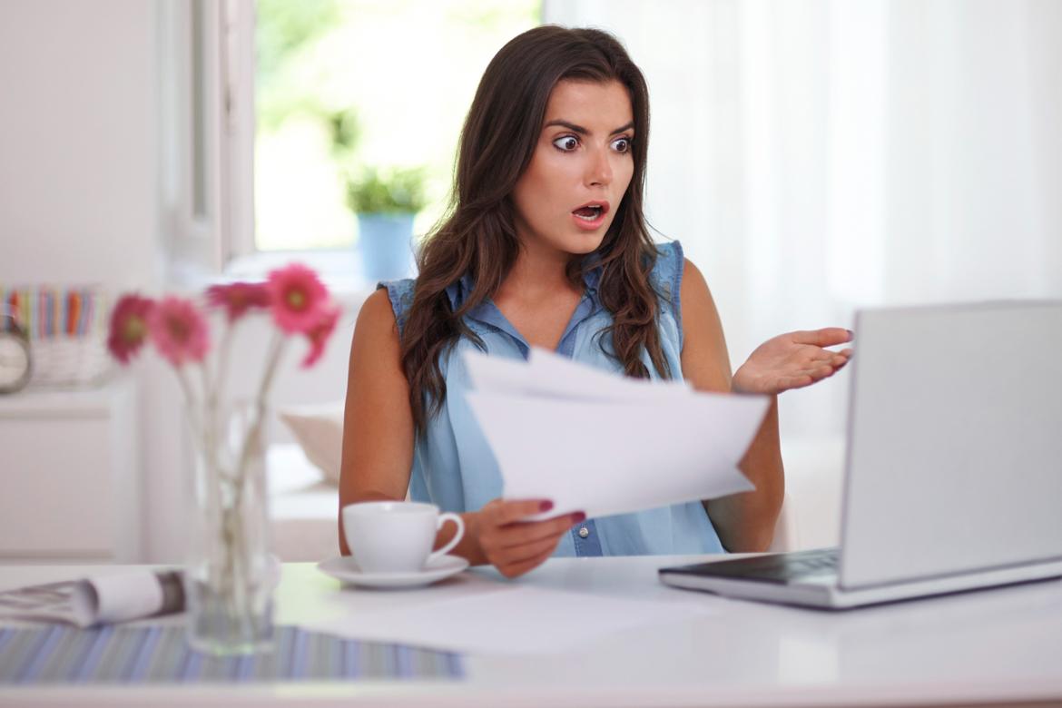 Woman at a desk holding papers and looking shocked at her laptop screen.