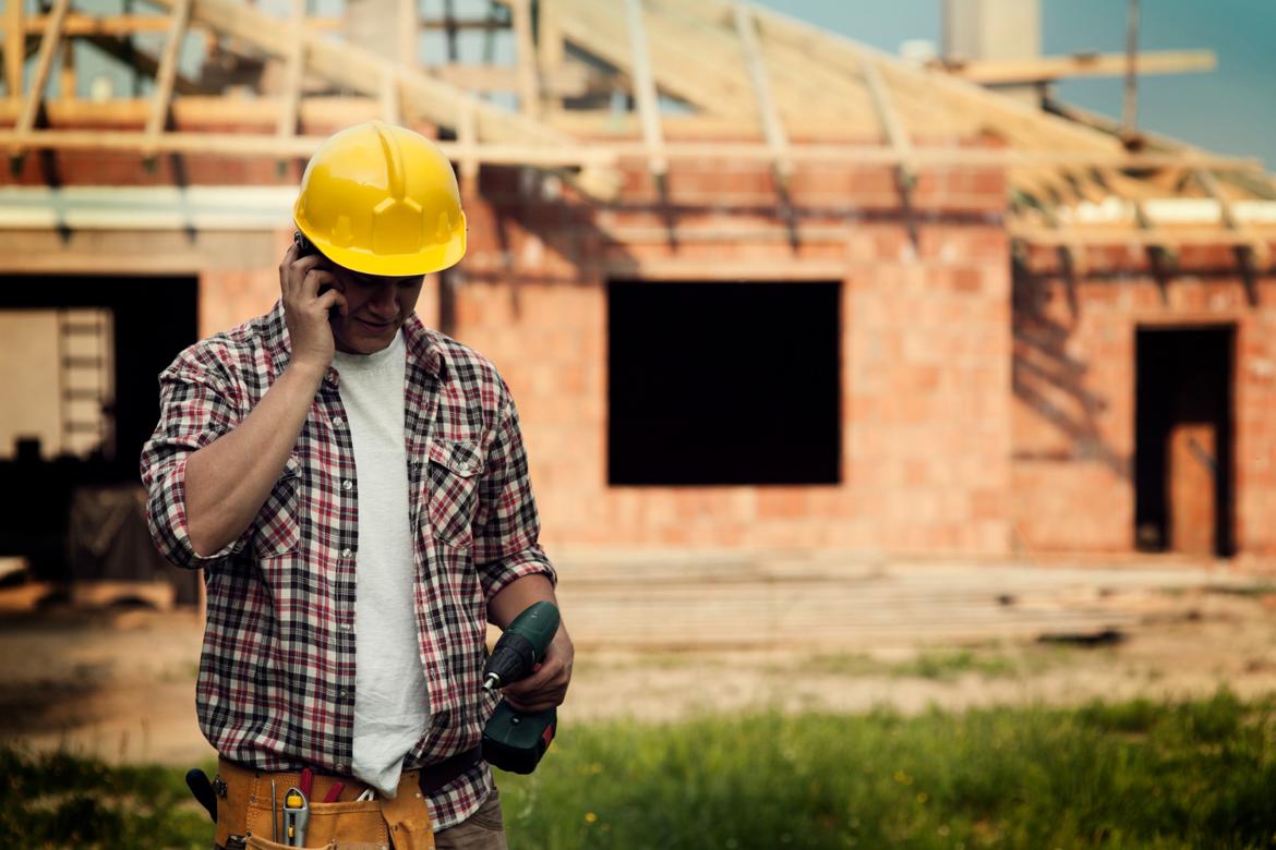 Man wearing a hard hat and toolbelt and holding a drill talking on a phone in front of a house under construction.