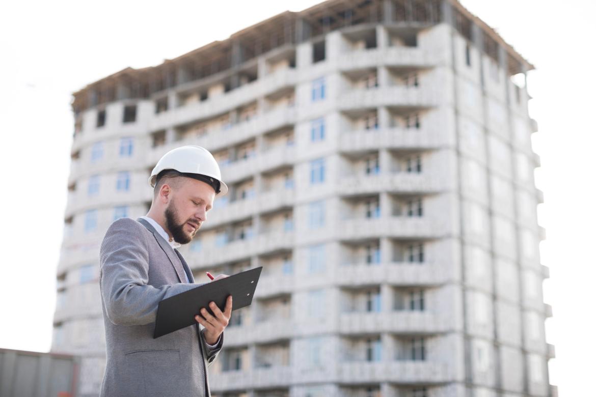 Young man in a business suit writing on a clipboard at a condo construction site.