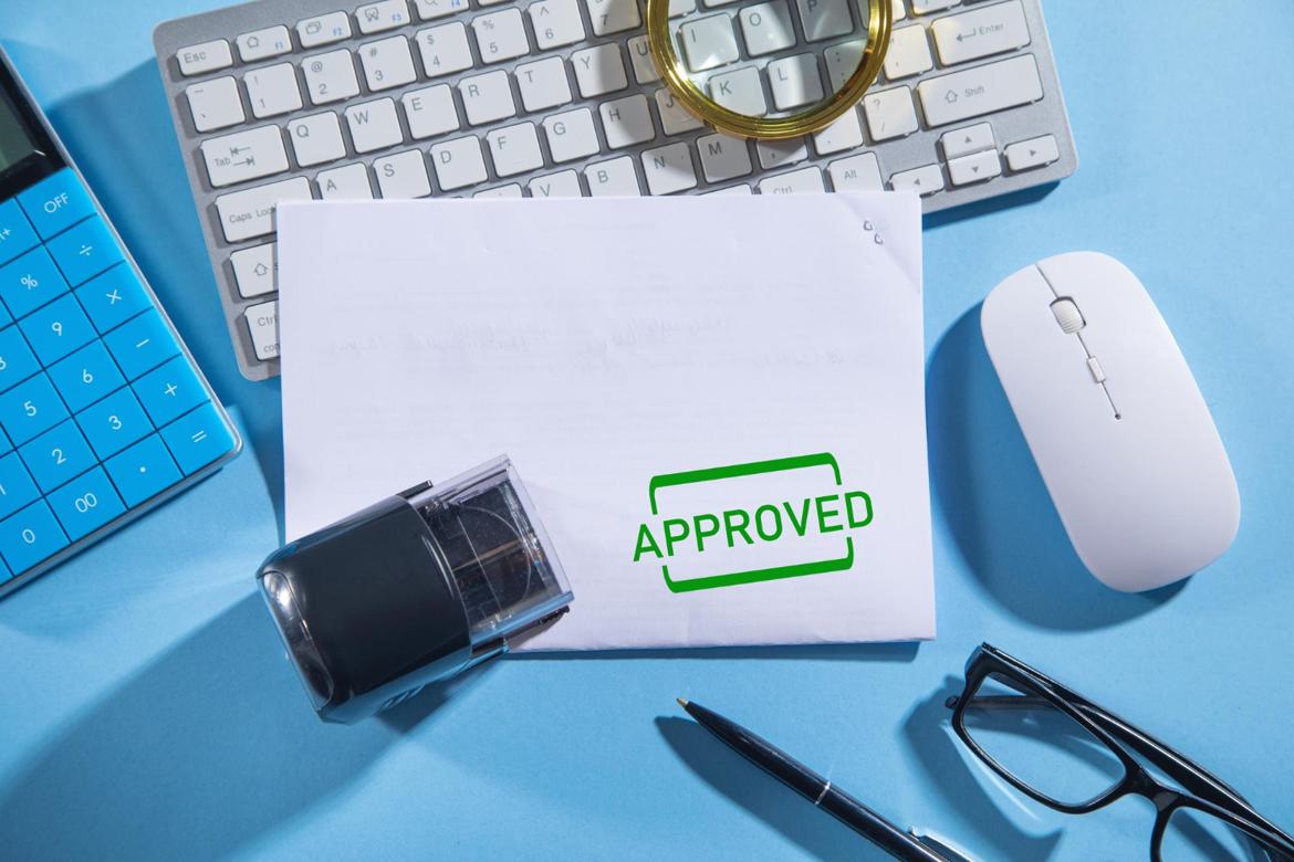 Document stamped approved on a desk with a keyboard and other business objects.